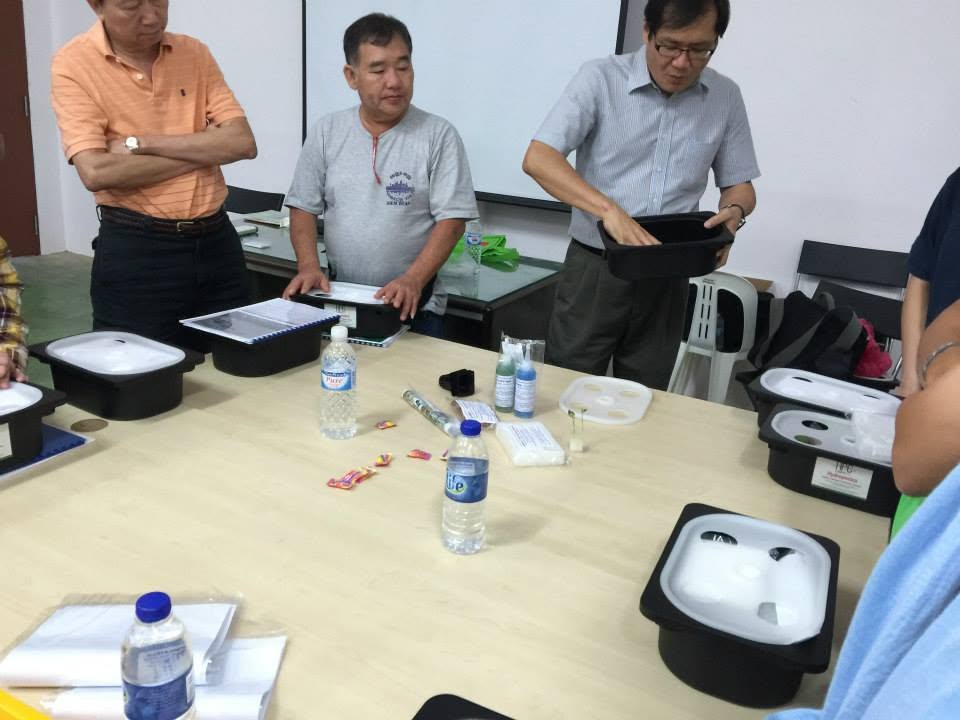 Participants standing around a table handling a black grow tub and its lid, with net pots, nutrient bottles and growing media laid out in front of them