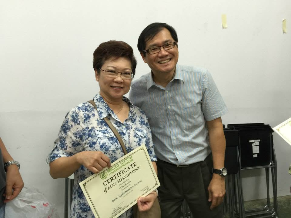 A smiling participant receiving her Certificate of Accomplishment for the Basic Hydroponics Course from the trainer at the end of the class