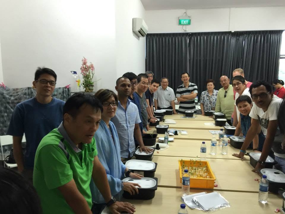 Participants seated along both sides of a long table, each with a black hydroponic grow tub and lid in front of them, turning to face the camera