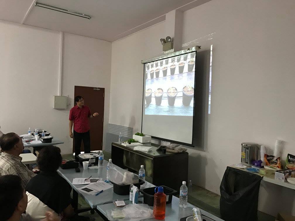 Geoffrey Chow presenting to the class beside a projected photograph of a row of pots with seedlings at different stages of germination