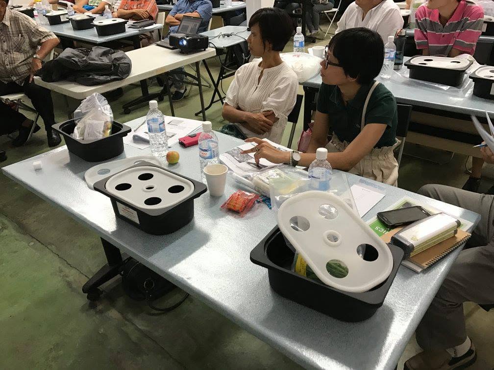 Hydroponic grow tubs with white lids on classroom tables, one with four planting holes and another with six, alongside notes and supplies