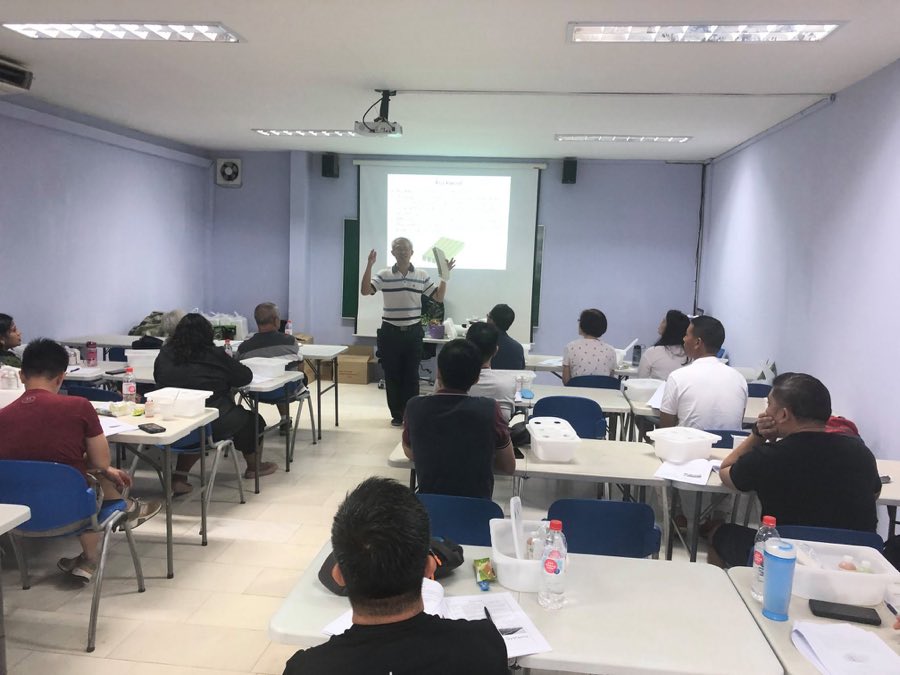 Trainer presenting hydroponic system types to seated learners in a classroom, each desk holding a white hydroponics starter kit