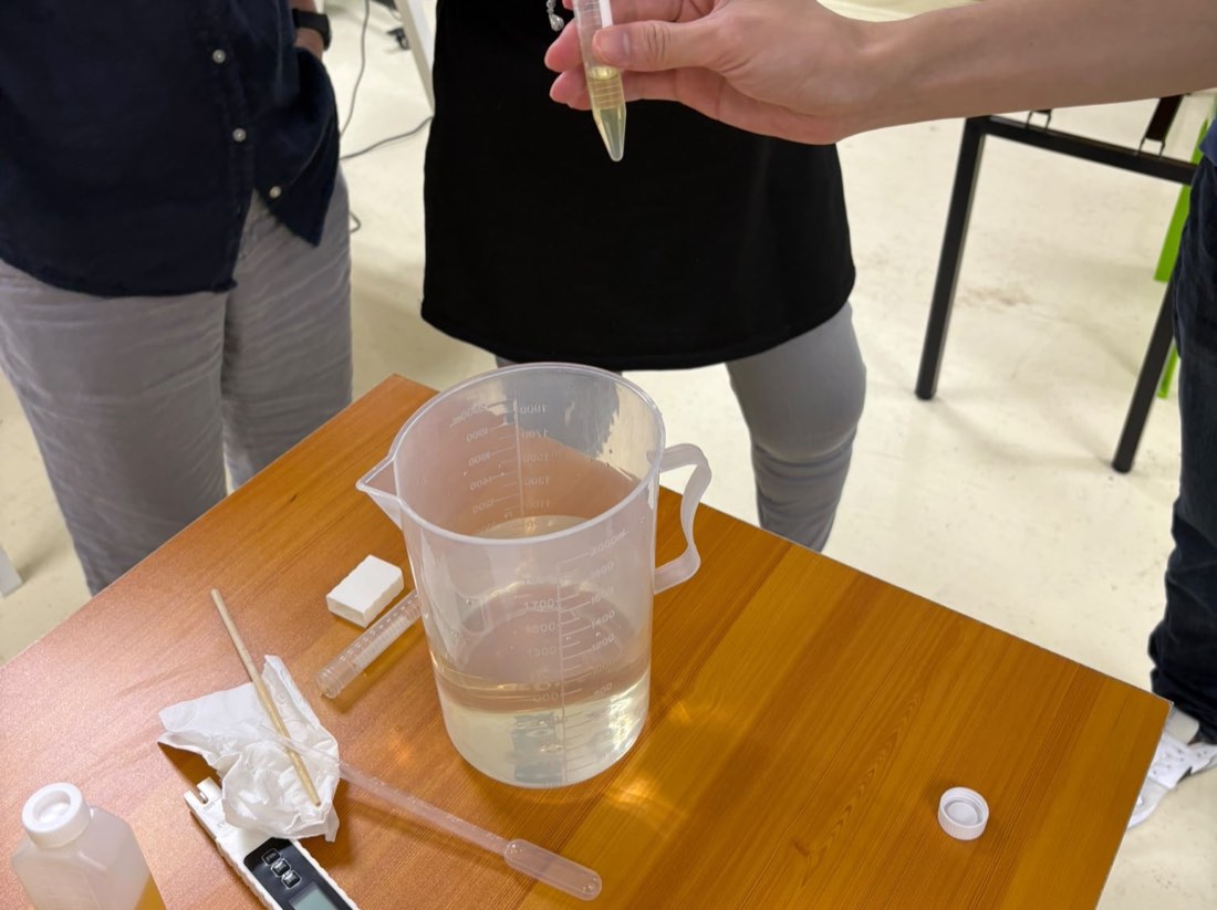 Class participant holding a test tube of concentrated hydroponic nutrient over a 2-litre beaker of mixed solution, with a TDS and EC meter, pipette and pH test strips on the bench