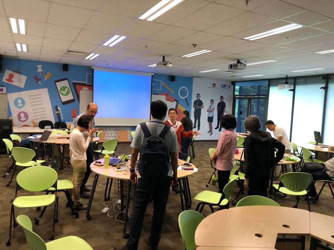 ITE staff standing and working at round tables with laptops and IoT components during the hands-on lab, projector screen lit at the front of the iROOM