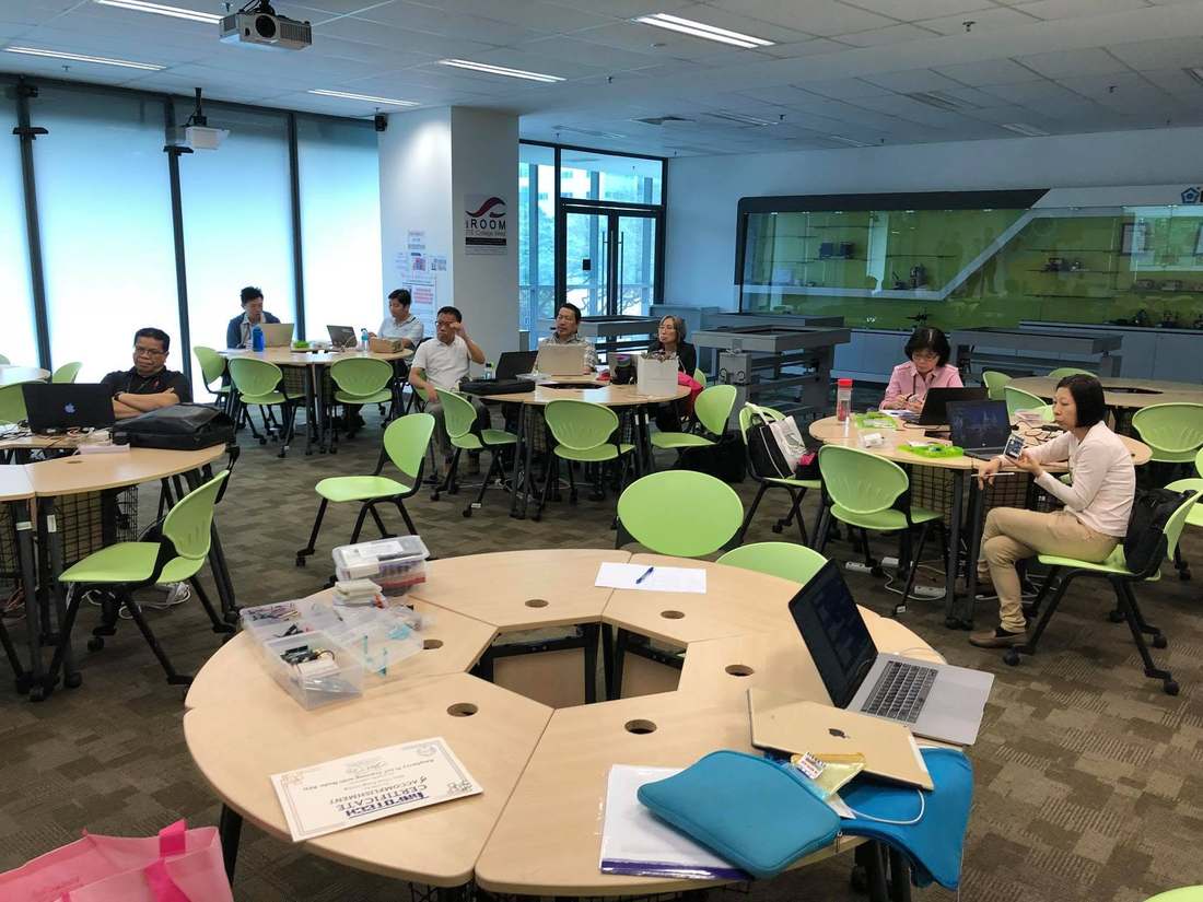 Wide view of the iROOM at ITE College West with staff at round tables, laptops open and boxes of IoT components and sensors on the foreground table