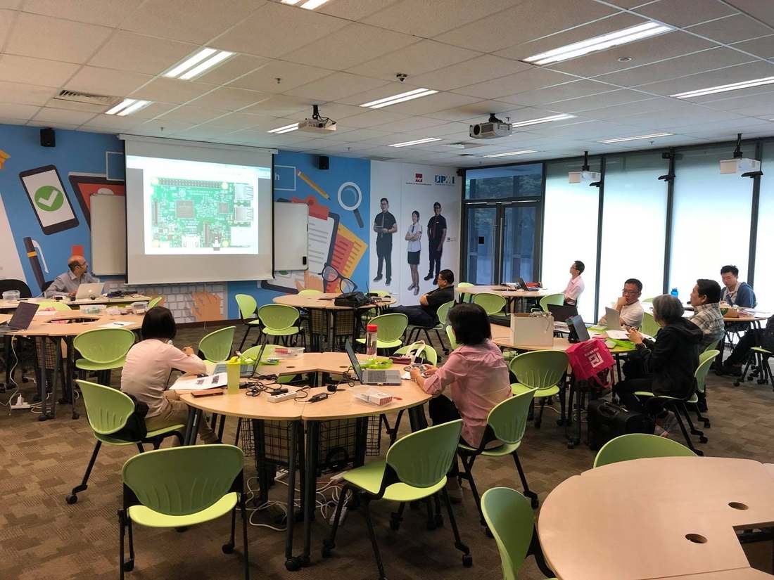 Trainer presenting a slide showing a Raspberry Pi board on the projector screen while ITE staff follow along at round tables with laptops
