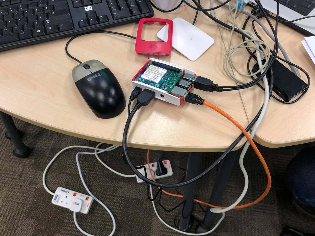A Raspberry Pi in a red and white case on a classroom desk, connected by USB and an orange Ethernet cable, next to a keyboard and mouse