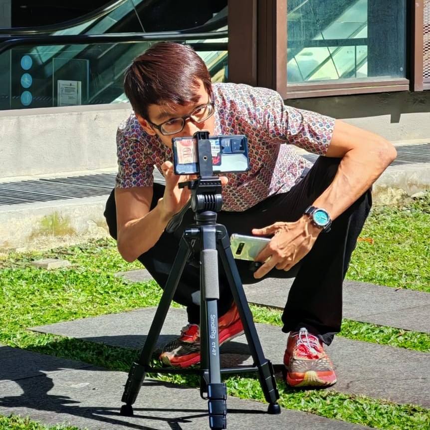 A learner crouched behind a smartphone mounted on a small tripod, composing a low-angle shot outdoors